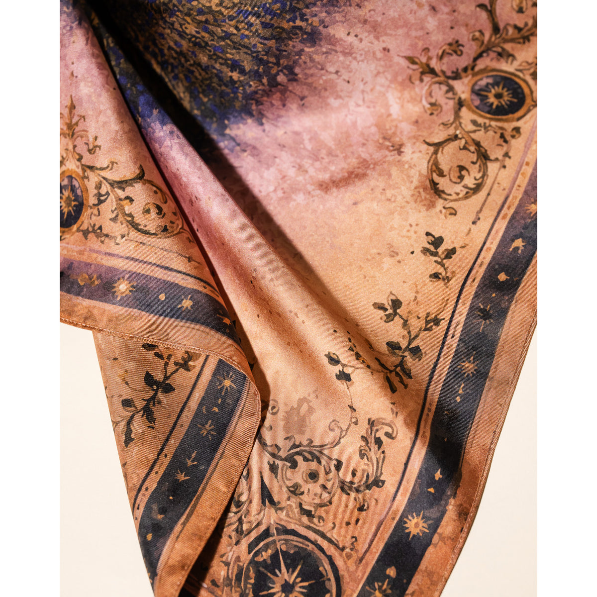 A close-up of a square scarf with a warm peach and beige base, featuring dark navy blue borders with gold star motifs and black botanical scrollwork.