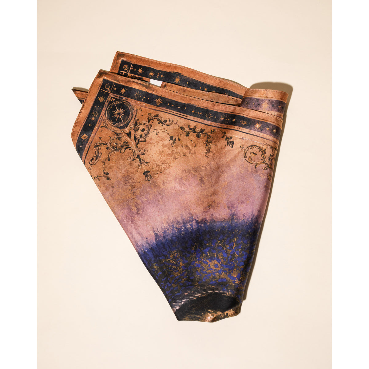 A folded silk scarf with a celestial pattern, featuring a gradient from copper-brown and lavender to a deep navy blue at the bottom, with gold star and moon details.