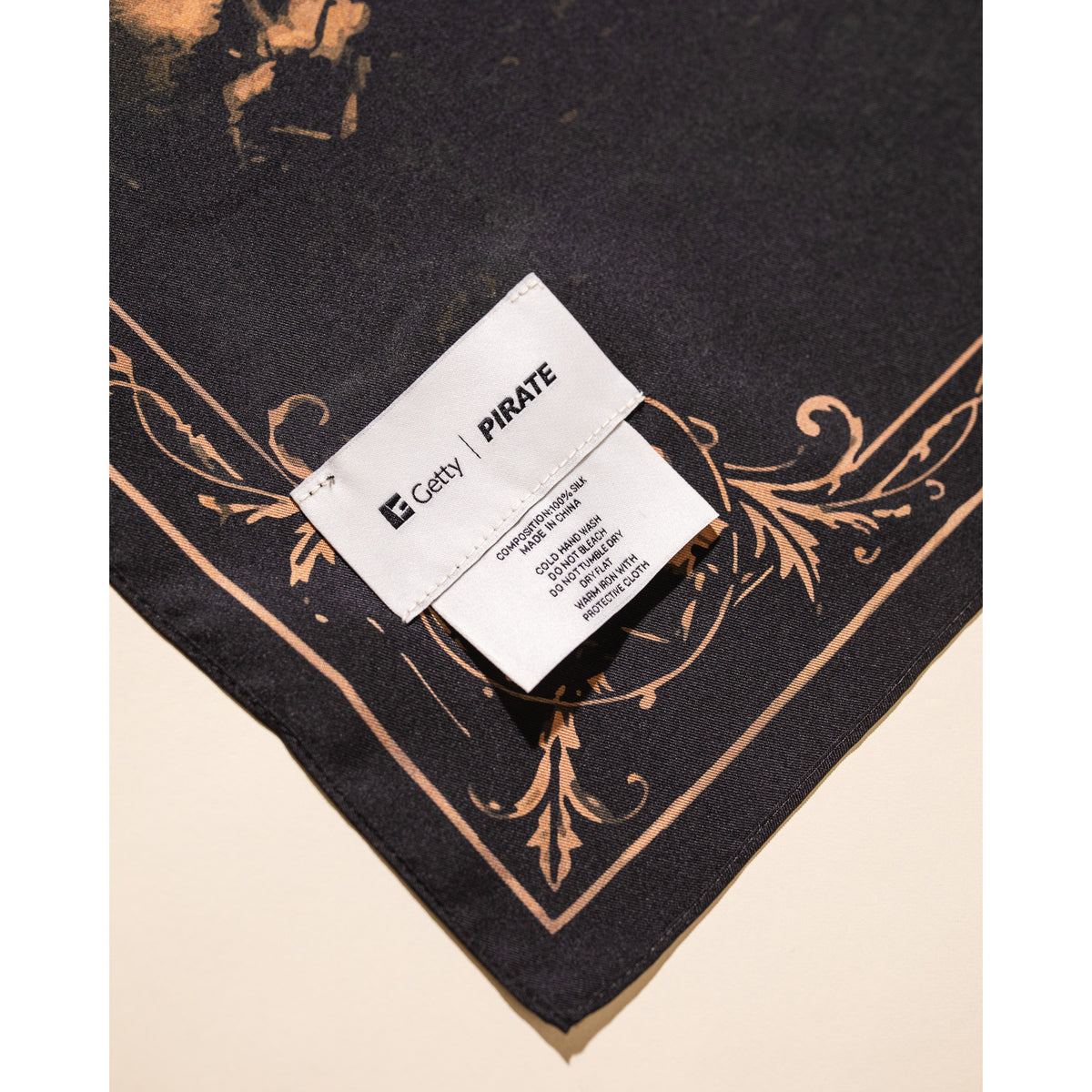 A close-up of a black scarf with a beige baroque-style border pattern, featuring a white sewn-in label that reads 'Getty | PIRATE'.