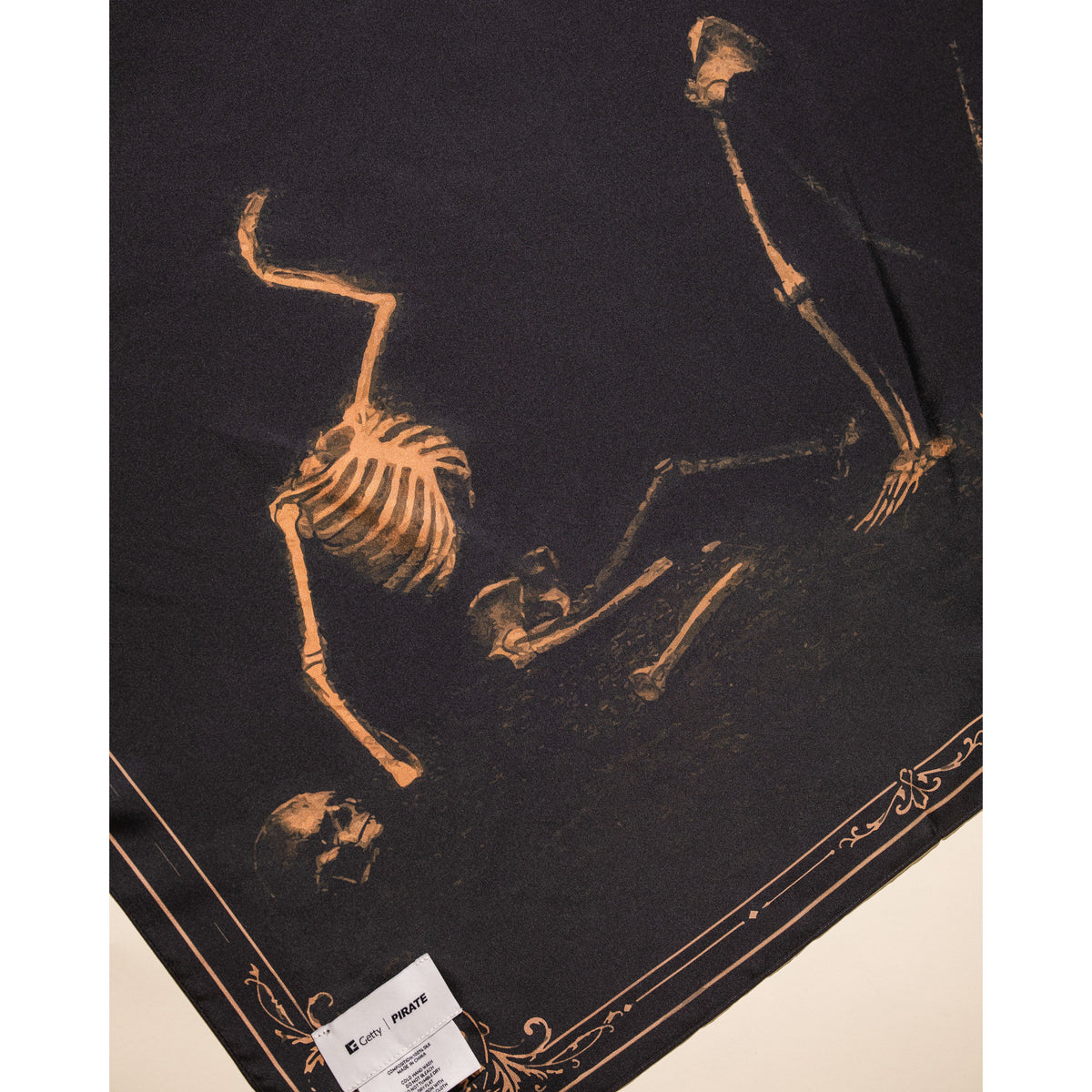 A black fabric item printed with a gothic-style skeleton design in a warm tan/gold color. The fabric is laid flat on a light beige surface, with a white paper tag visible in the bottom-left corner.