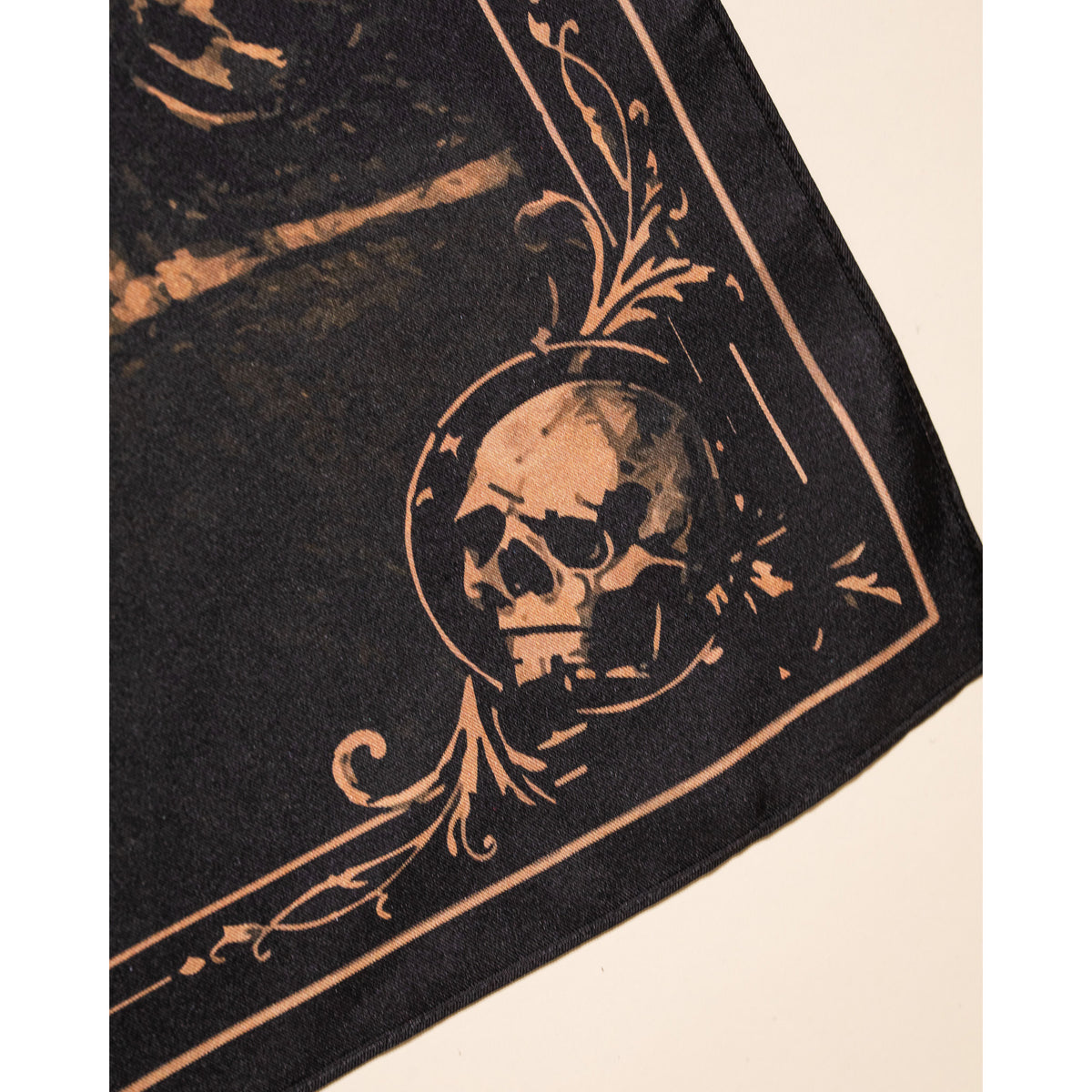 A close-up of a black fabric product, likely a scarf or bandana, featuring a skull and ornate baroque scrollwork printed in a muted tan or beige color.