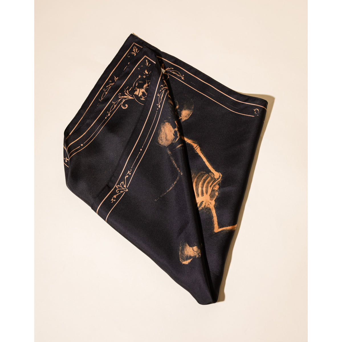 A black silk scarf with a gold skeleton and decorative border print, folded diagonally on a light beige background.