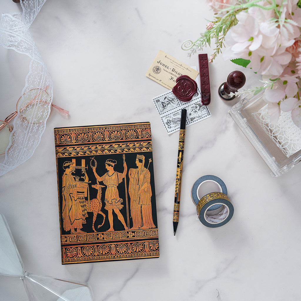 A styled flat-lay featuring a black and gold ancient Greek-style notebook or card with a mythological illustration, surrounded by wax seals, a pen, washi tape, and decorative props on a white marble surface.
