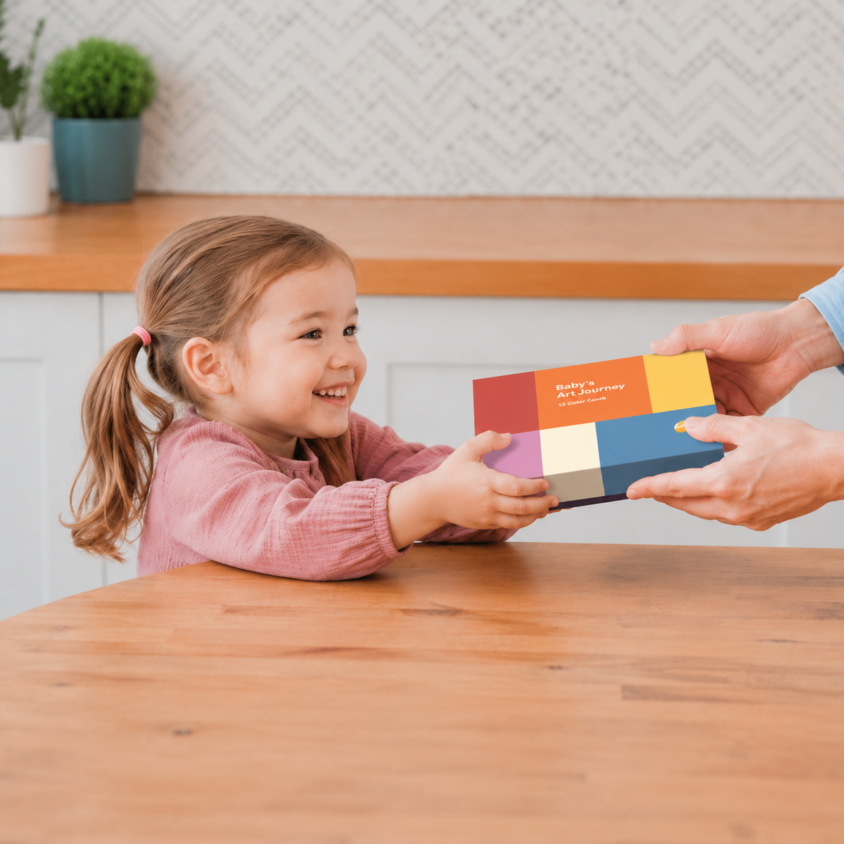 A young girl smiling as an adult hands a colorful rectangular card or box to her at a wooden table in a bright kitchen.