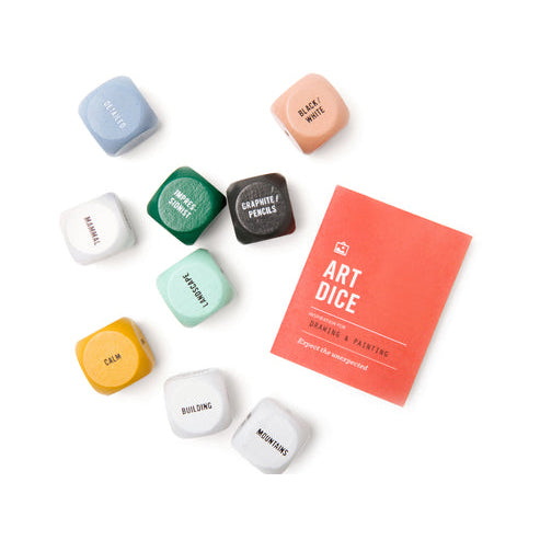 A set of colorful art dice for designers and painters, shown with a coral orange instruction card on a clean white background. The dice are labeled with terms like 'BRUSHSTrokes', 'COMBINE COLORS', 'BLACK/WHITE', 'MATERIAL', 'JEWELRY', 'CASH', 'BUILDING', and 'MOTIONS'.