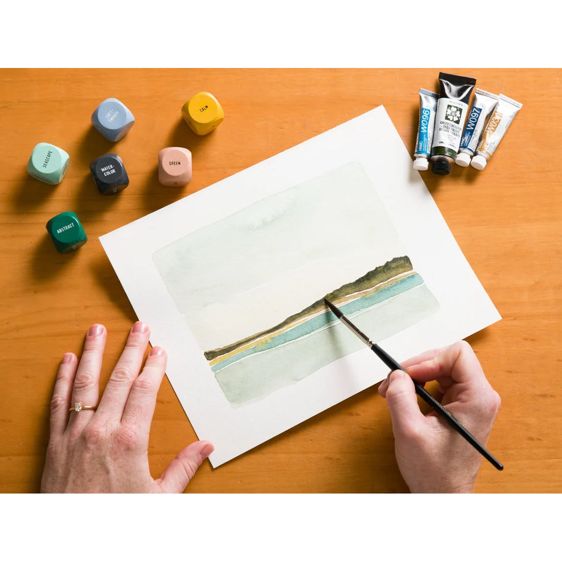 A top-down view of a person's hands painting on a sheet of white watercolor paper with a landscape scene. The paper is placed on a wooden table, surrounded by paint cubes and tubes of paint.