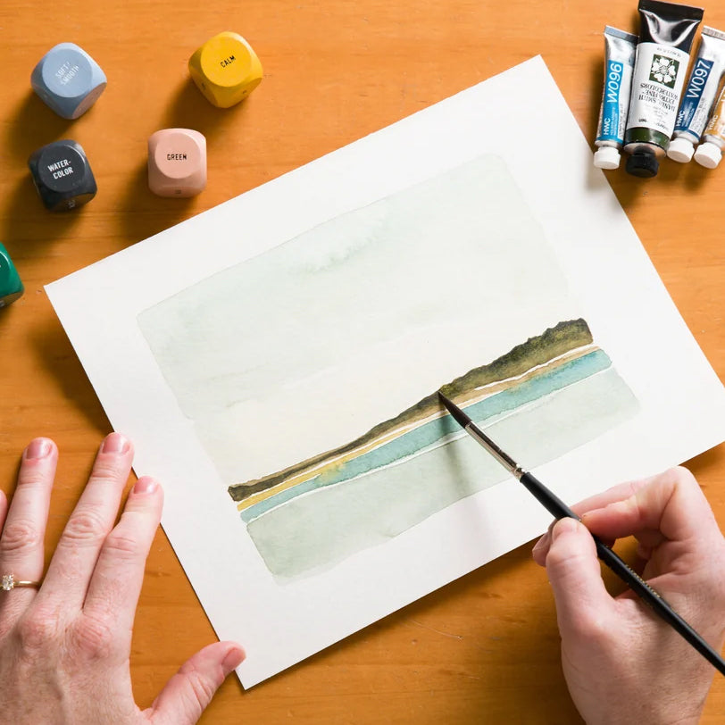 A watercolor landscape painting in progress on white paper, with a hand holding a paintbrush applying strokes to the lower right. Surrounding the artwork are paint cubes, tubes of acrylic paint, and the artist's hands on a wooden table.