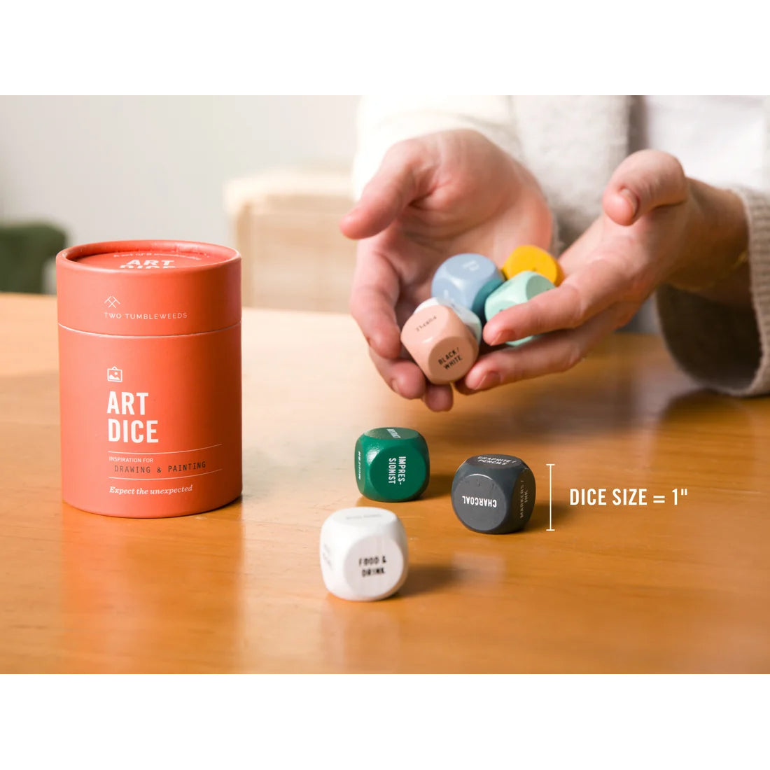 A person holds several small colorful art dice over a wooden table next to a round orange container labeled ART DICE. Additional dice are placed on the table, and a size marker reads 'DICE SIZE = 1"'.