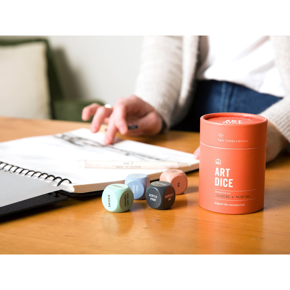 A person seated at a wooden table with an open notebook in a real indoor setting, with several small colored dice and a cylindrical orange container labeled ART DICE on the table.