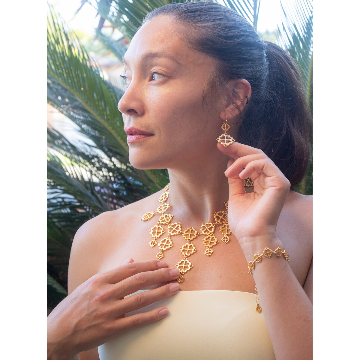 model wearing matching gold earrings, statement necklace and bracelet against a leafy backdrop 