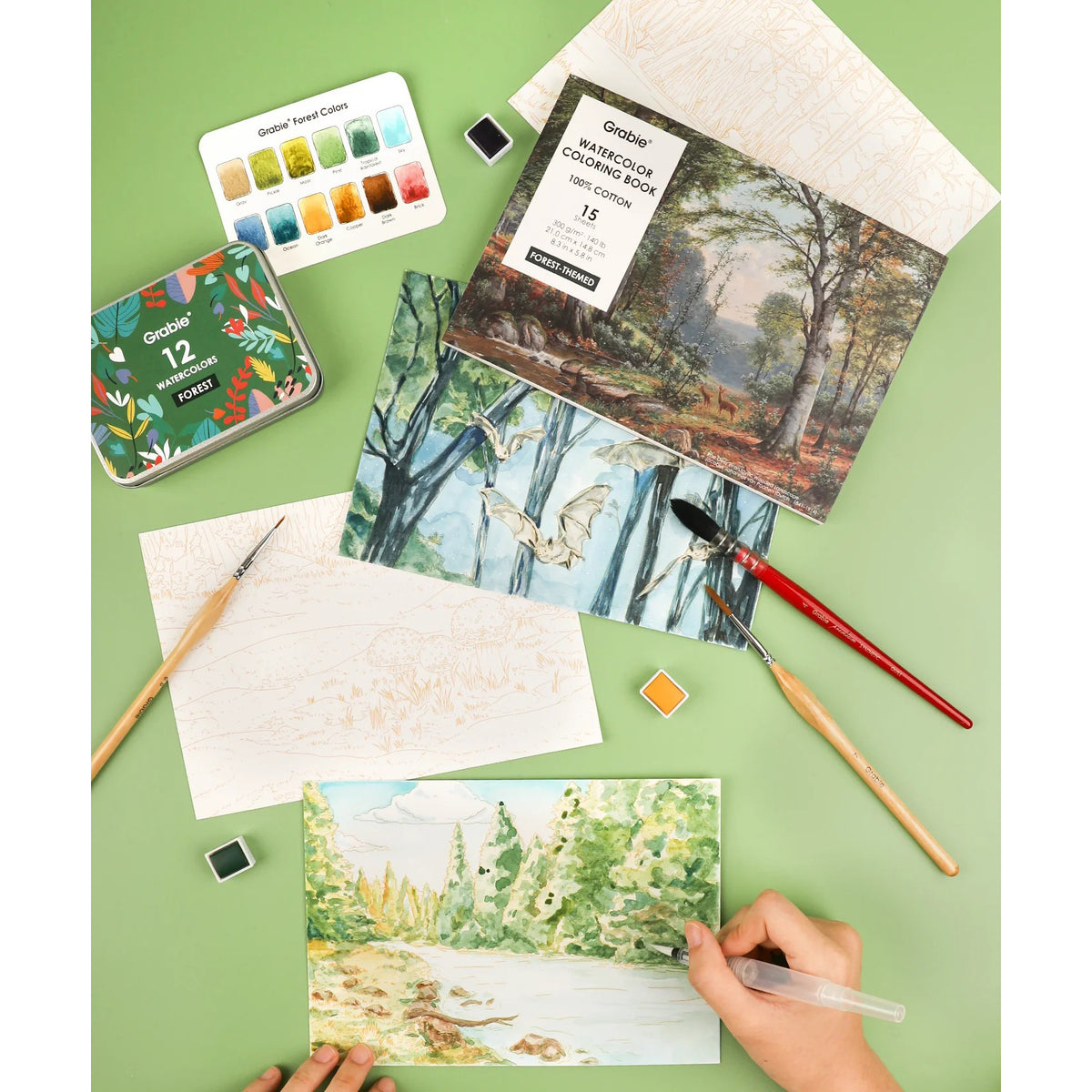 Art supplies including watercolor paint sets, brushes, and a painting of a forest scene on a green surface.