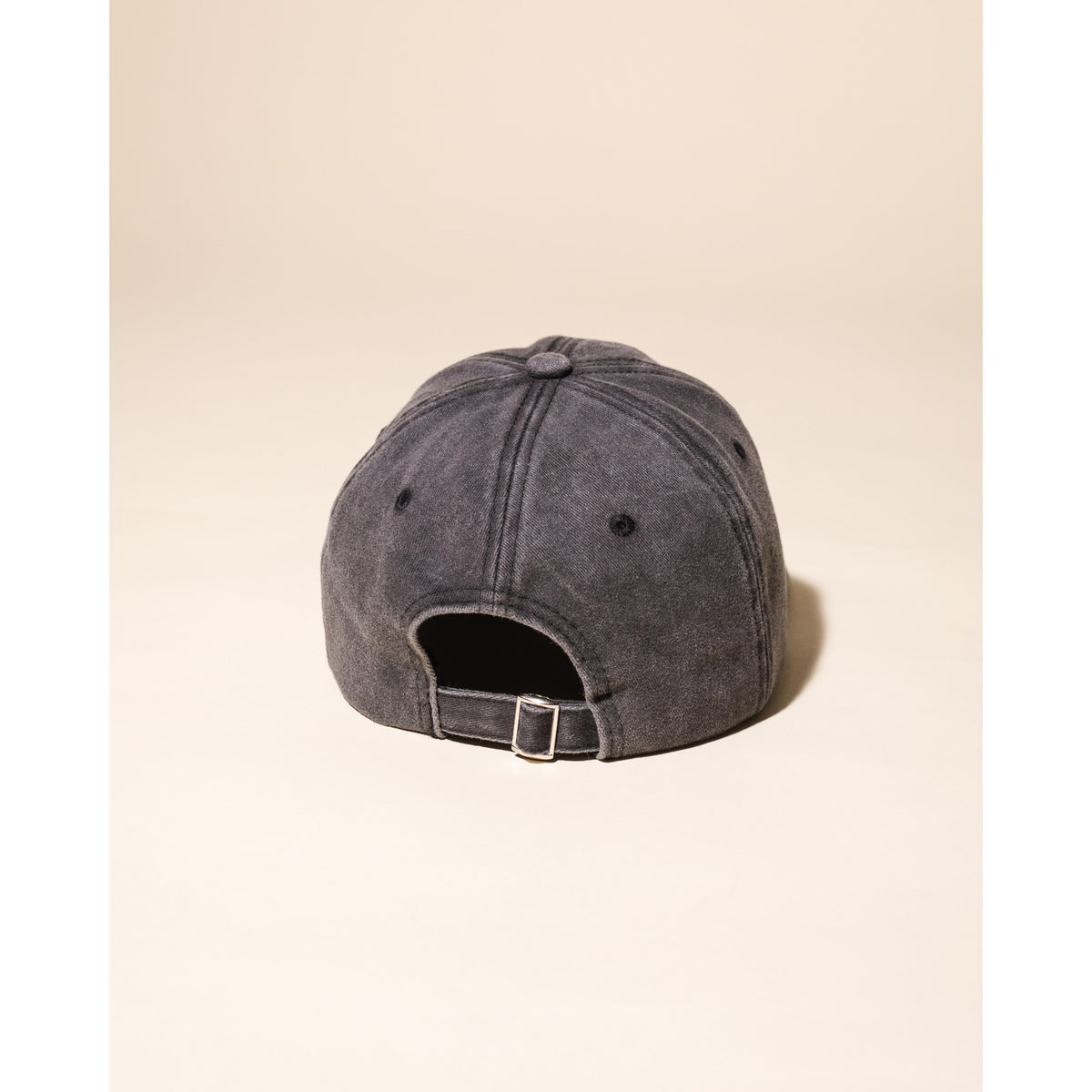 A washed charcoal gray baseball cap shown from the back, featuring a fabric strap and metal buckle closure, set against a solid beige background.