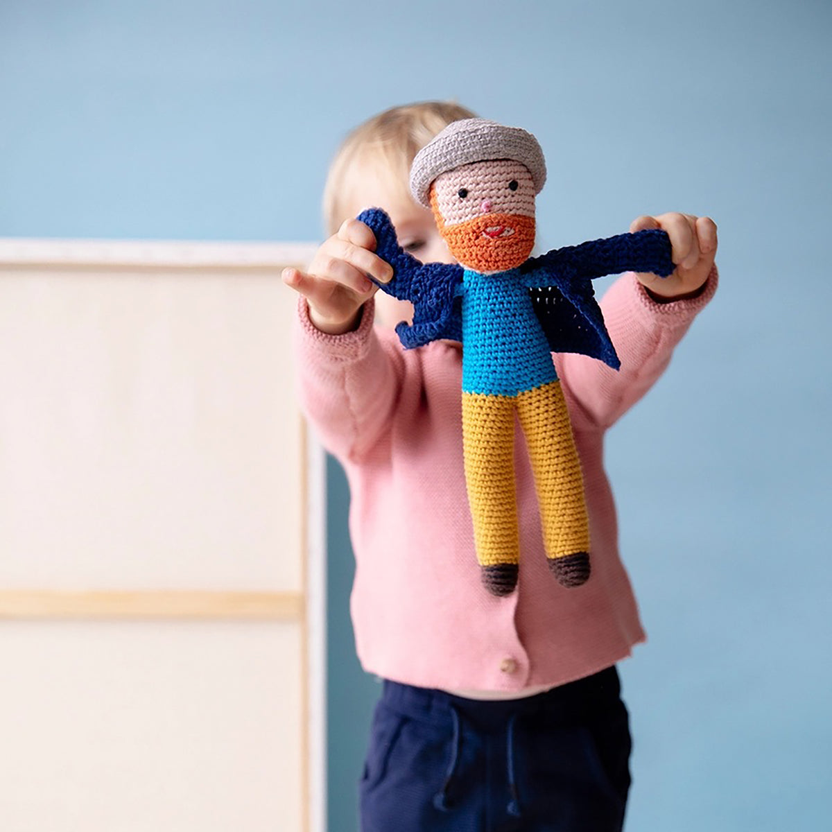 Child holding a crocheted doll with a blue outfit against a light blue wall.