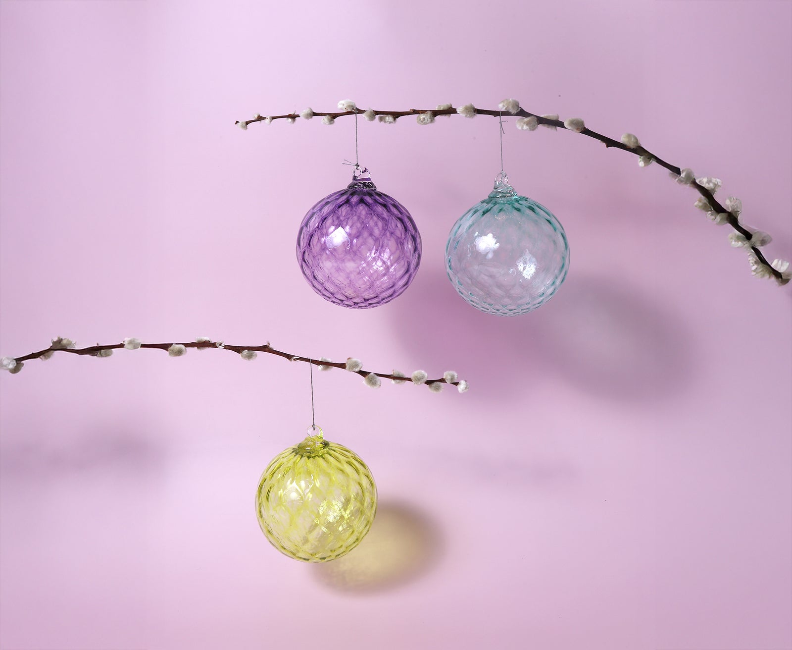 Three colorful glass ornaments hanging from branches against a pink background