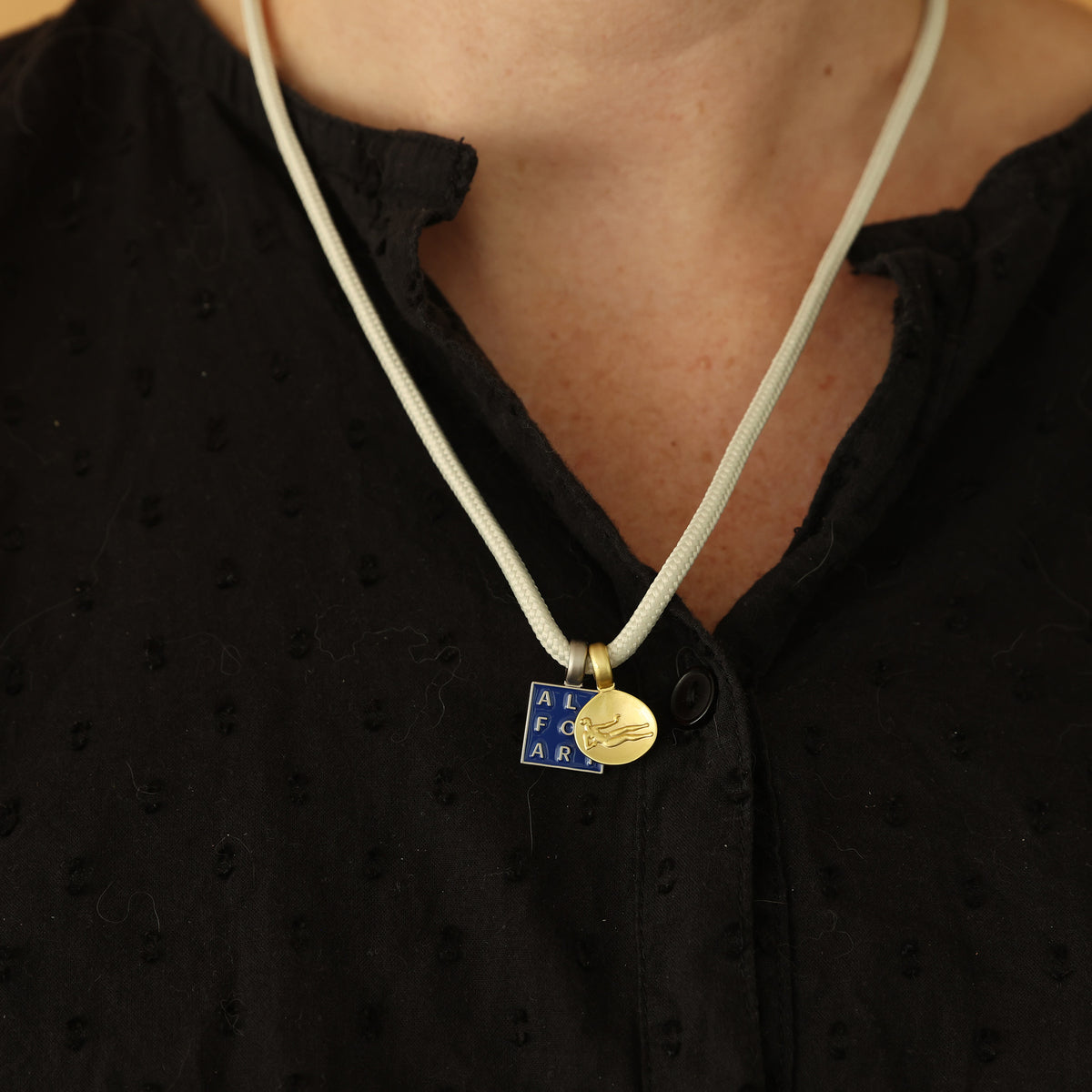 A close-up of a person wearing a white cord necklace with two pendants: a blue and white square enamel block reading 'ALL FOR ART' and a small gold disc charm.