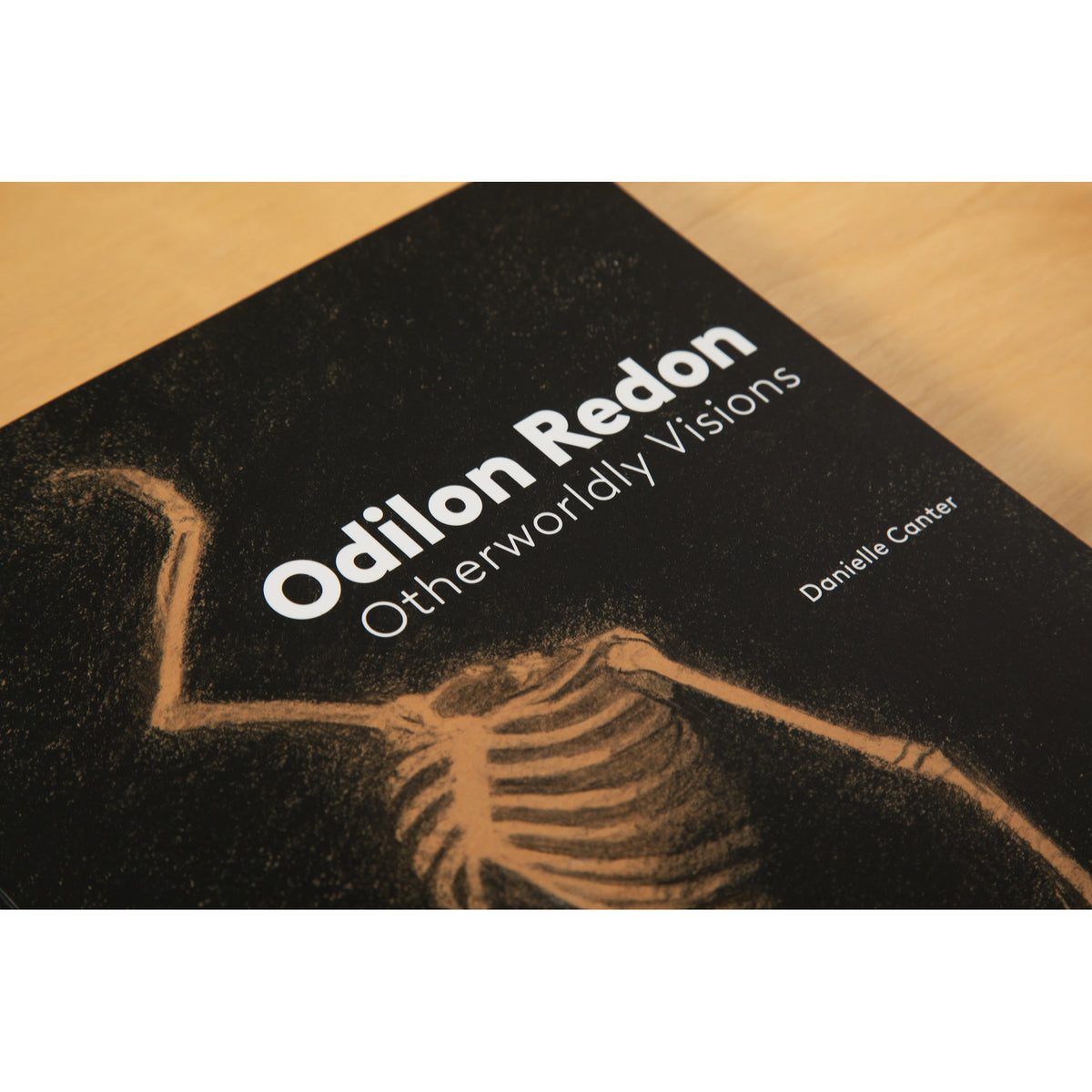 A book cover titled 'Odilon Redon: Otherworldly Visions'  featuring a black background with a tan skeleton illustration and white text, photographed on a light wood surface.