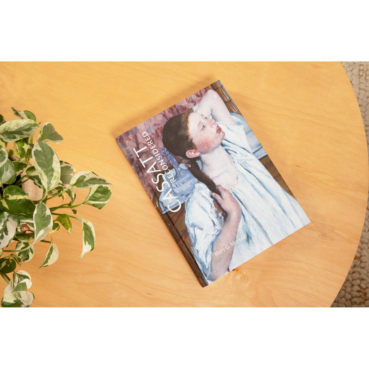 A book lies on a light wood table next to a green variegated plant.