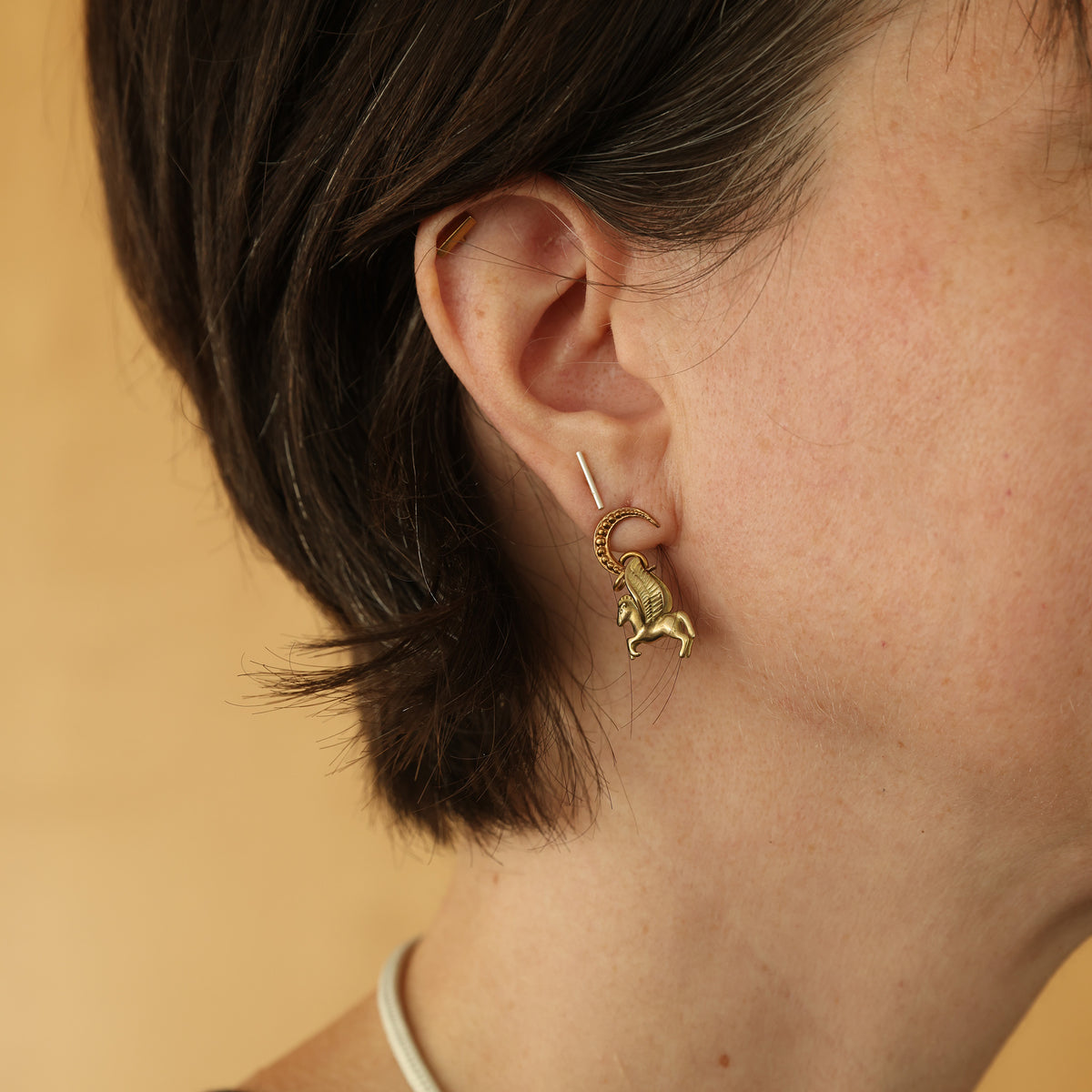 A close-up of a person's ear wearing a small gold earring with a dangling horse charm and a matching curved gold ring.