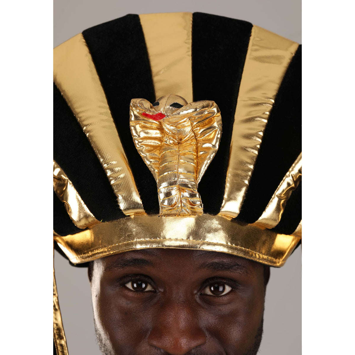 A close-up of a person wearing a shiny gold and black striped pharaoh-style headdress with a cobra motif on the front, against a plain gray background.