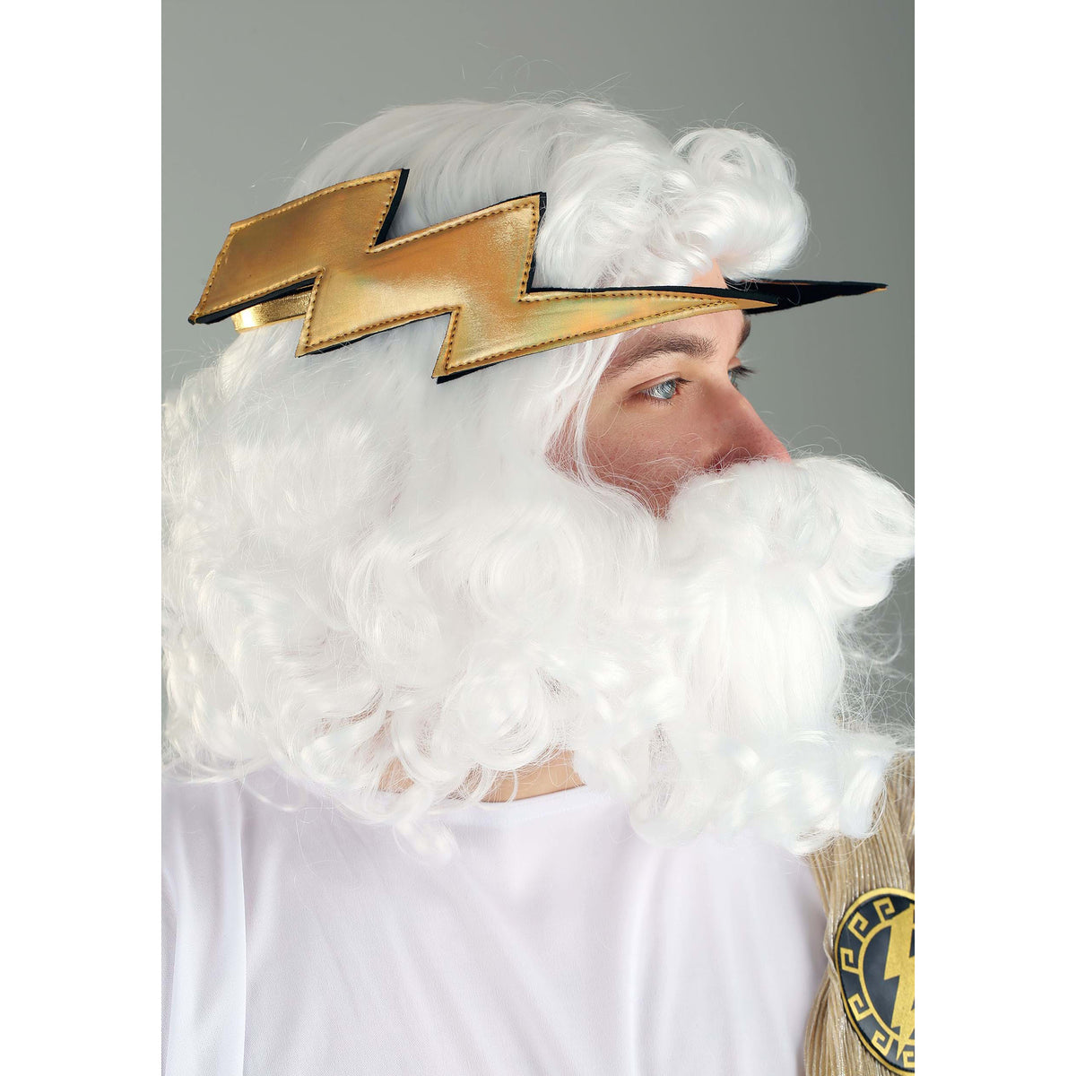 A man wearing a white curly wig, a white beard, and a gold lightning bolt headpiece against a plain gray background.