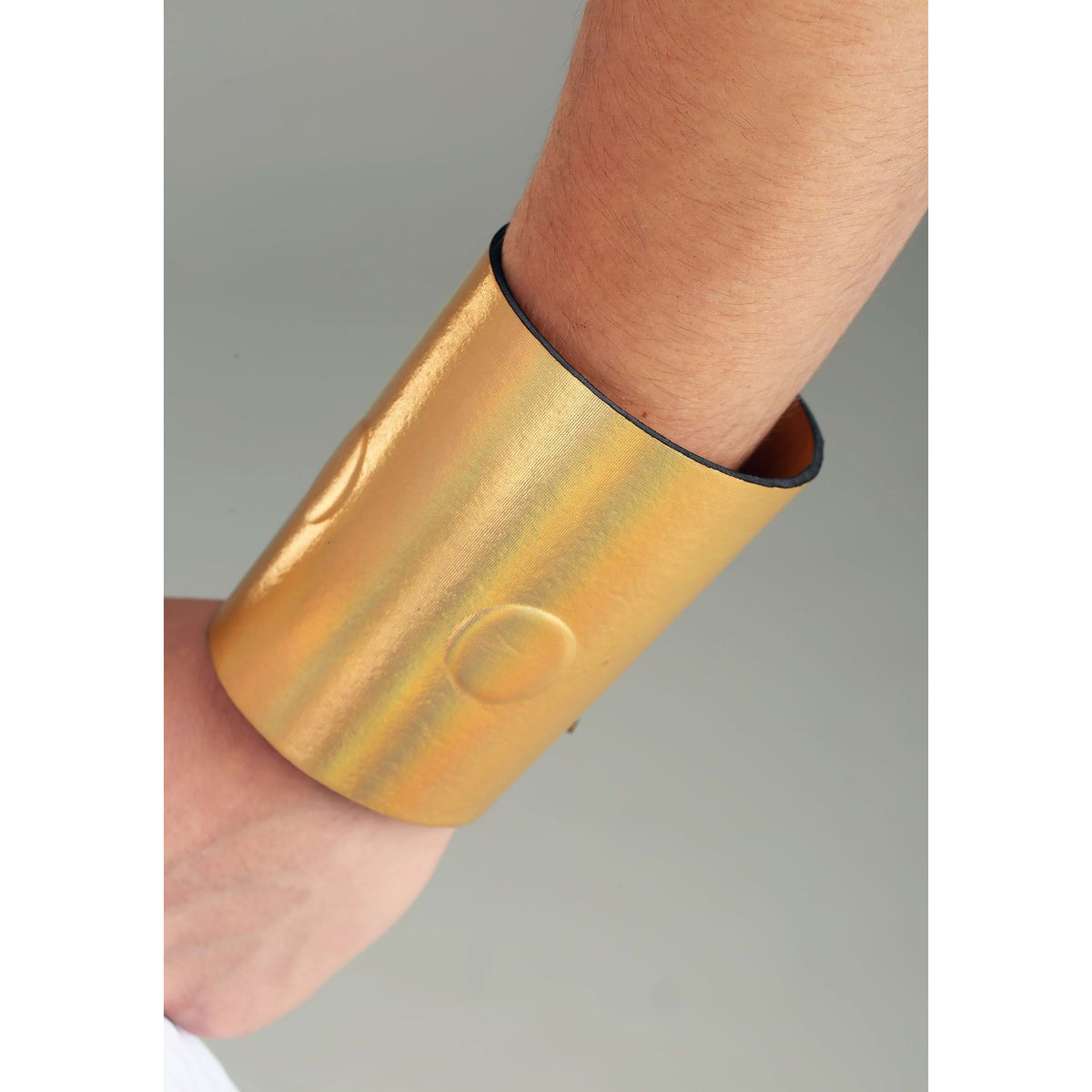 A close-up of a person's forearm and hand wearing a wide, metallic gold cuff bracelet against a neutral gray background.