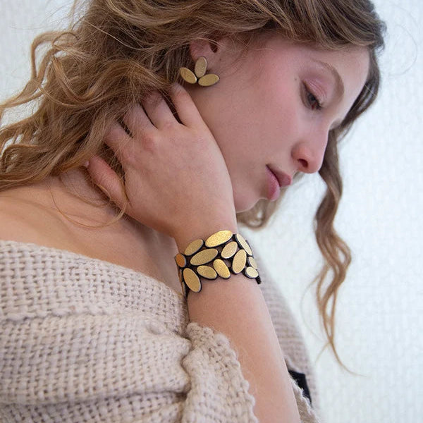 A woman wearing a chunky gold leaf-patterned cuff bracelet and matching gold leaf earrings, styled with a cream knit sweater against a light textured wall.