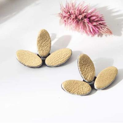 Three pairs of textured gold oval stud earrings arranged on a white surface with decorative pink dried flowers in the background.