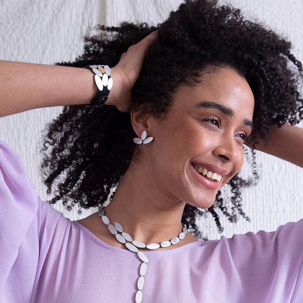 A smiling woman with curly black hair wearing a lavender top, a white shell-style necklace, matching white earrings, and a white bracelet, posing against a light textured wall.