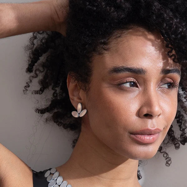 A close-up portrait of a woman wearing a black sleeveless top covered in layered silver oval shell-like embellishments, with matching silver earrings, against a plain light background.