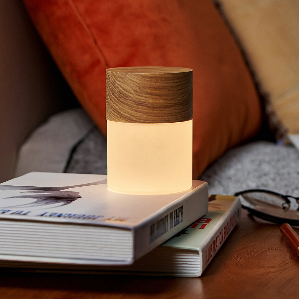 A small cylindrical bedside lamp with a glowing frosted lower half and a wood-grain top, resting on stacked books in a cozy indoor setting.