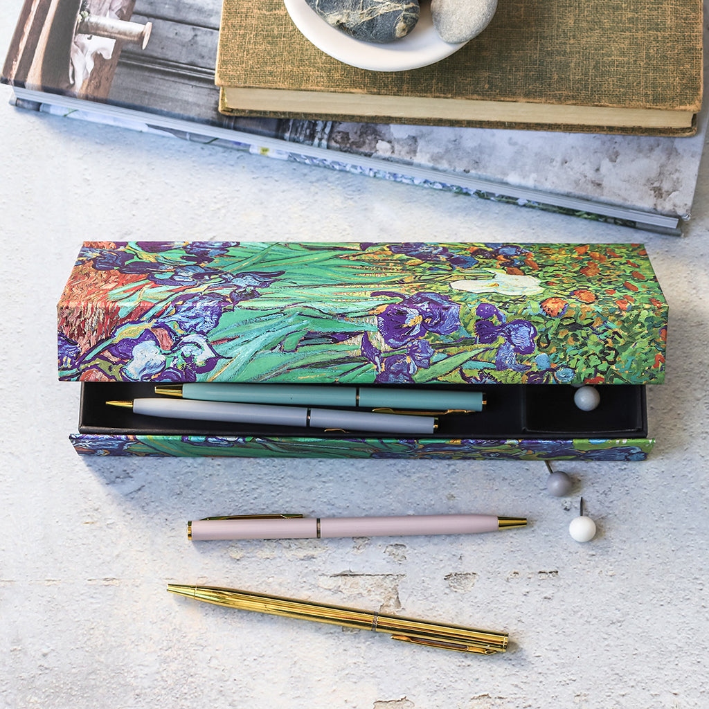 A decorative rectangular pen case with a floral iris print in green, purple, and blue, shown open with several pens inside. The case sits on a light textured surface with additional pens and books nearby.