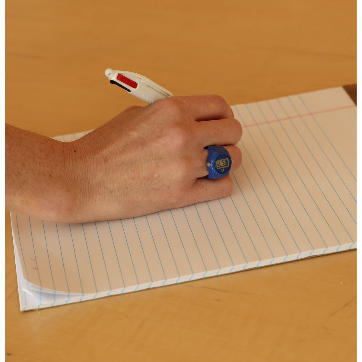 A close-up of a hand holding a white pen over lined notebook paper, wearing a blue ring-shaped novelty item on one finger.