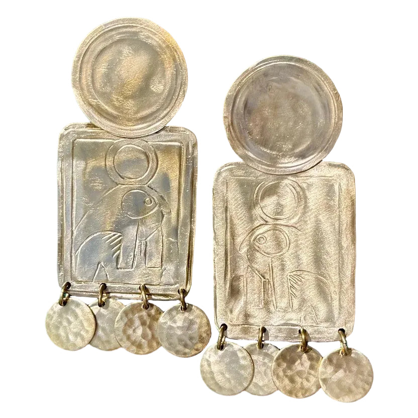 A pair of chunky, handcrafted silver-tone earrings with embossed figures and circular hammered dangles, displayed on a white background.