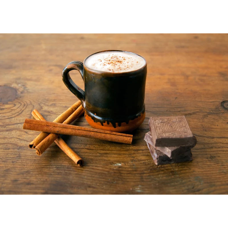 A dark glazed ceramic mug filled with a frothy hot drink, set on a rustic wooden table with cinnamon sticks and chocolate blocks nearby.