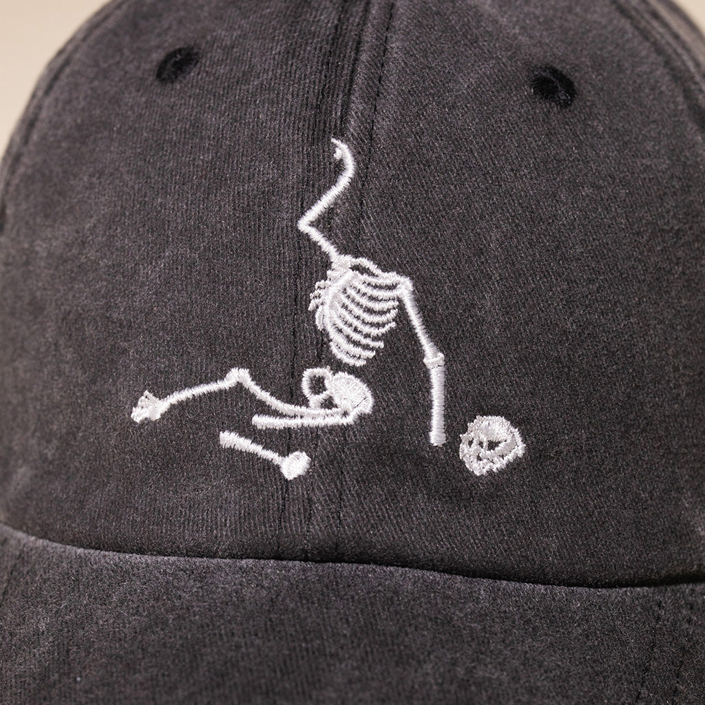 A close-up of a dark gray fabric baseball cap featuring a white embroidered skeleton 
