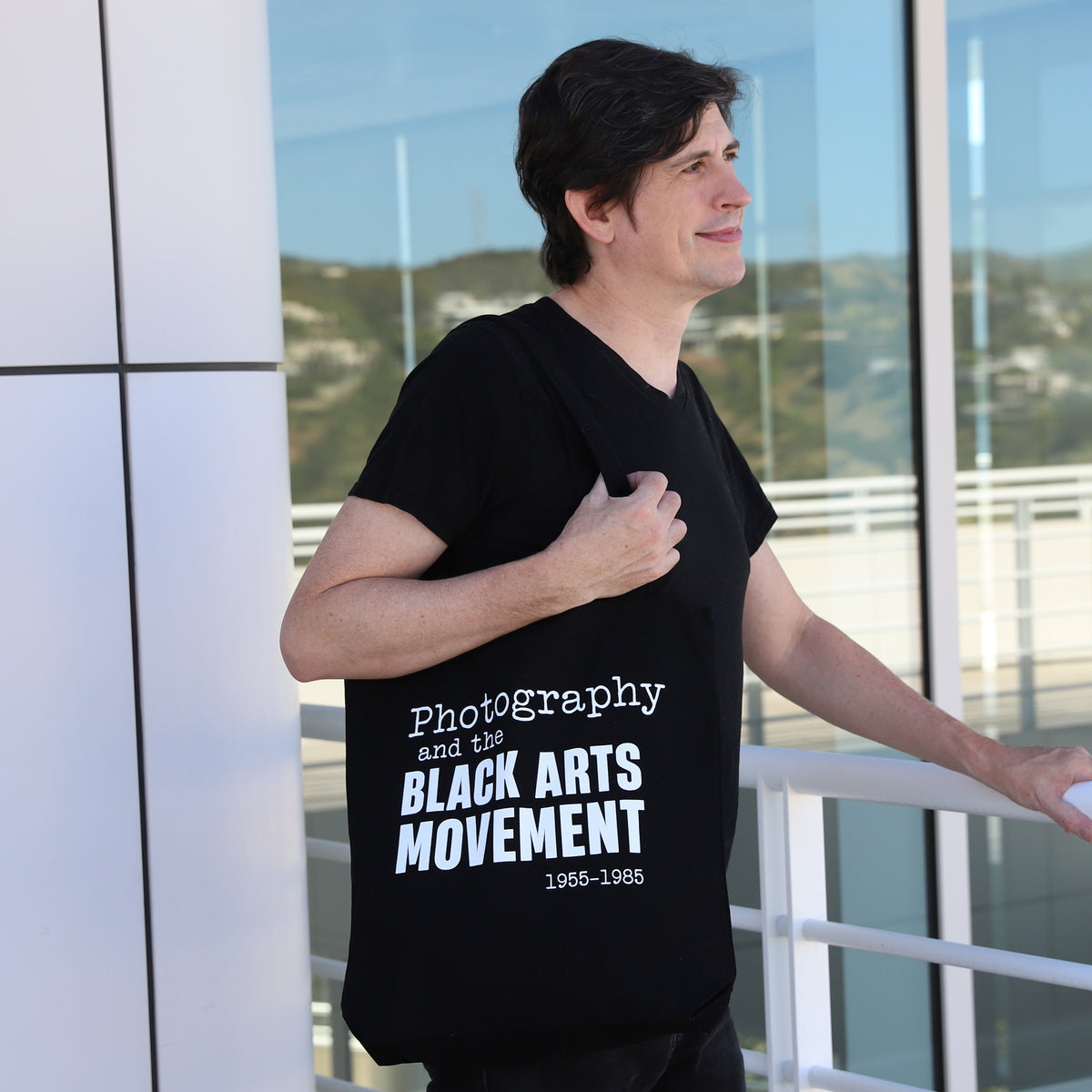 Man wearing a black t-shirt with text standing outdoors near a glass building.