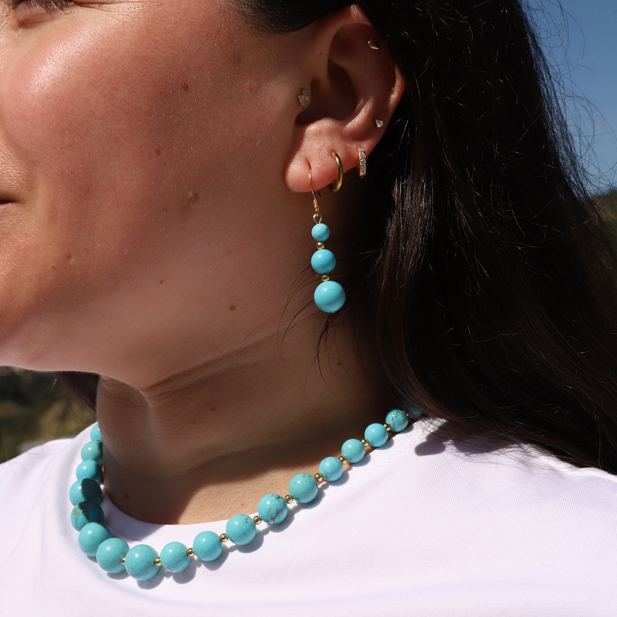Close-up of a person wearing turquoise beaded earrings and a necklace with a blurred natural background.