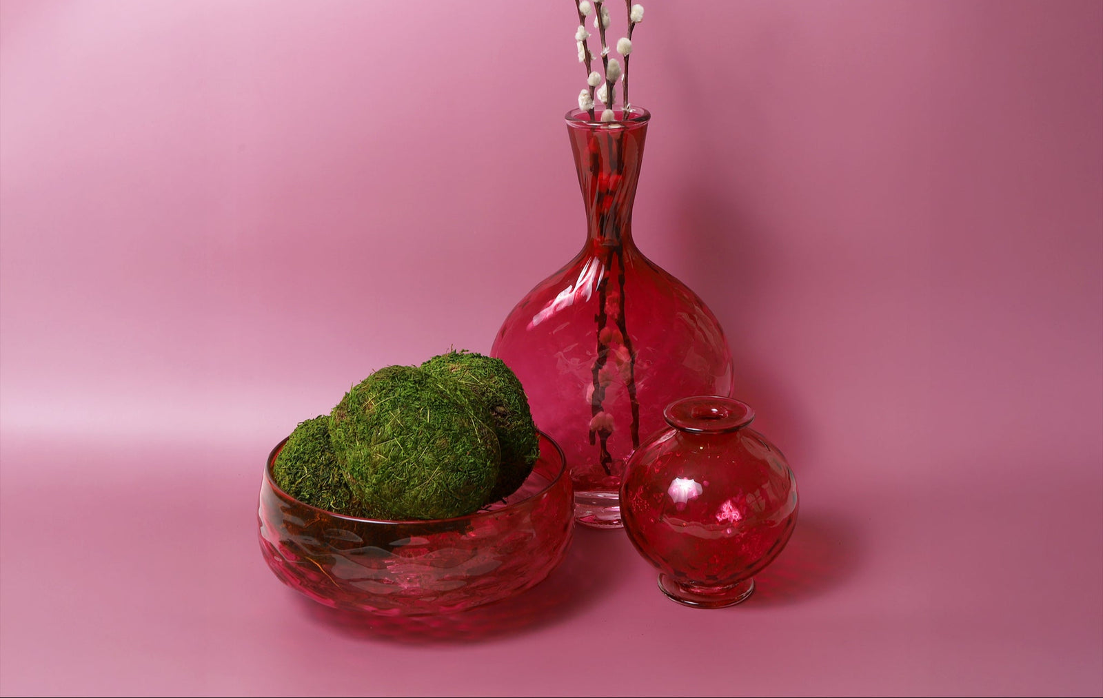 Three red glass vases with green moss and white branches on a pink background