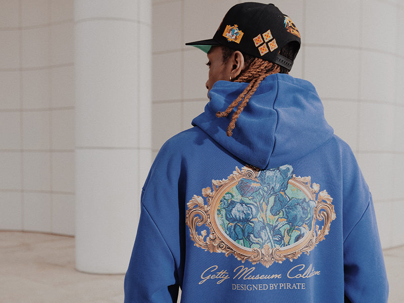 Person wearing a blue hoodie with a decorative back design and cap, standing against a white tiled wall.