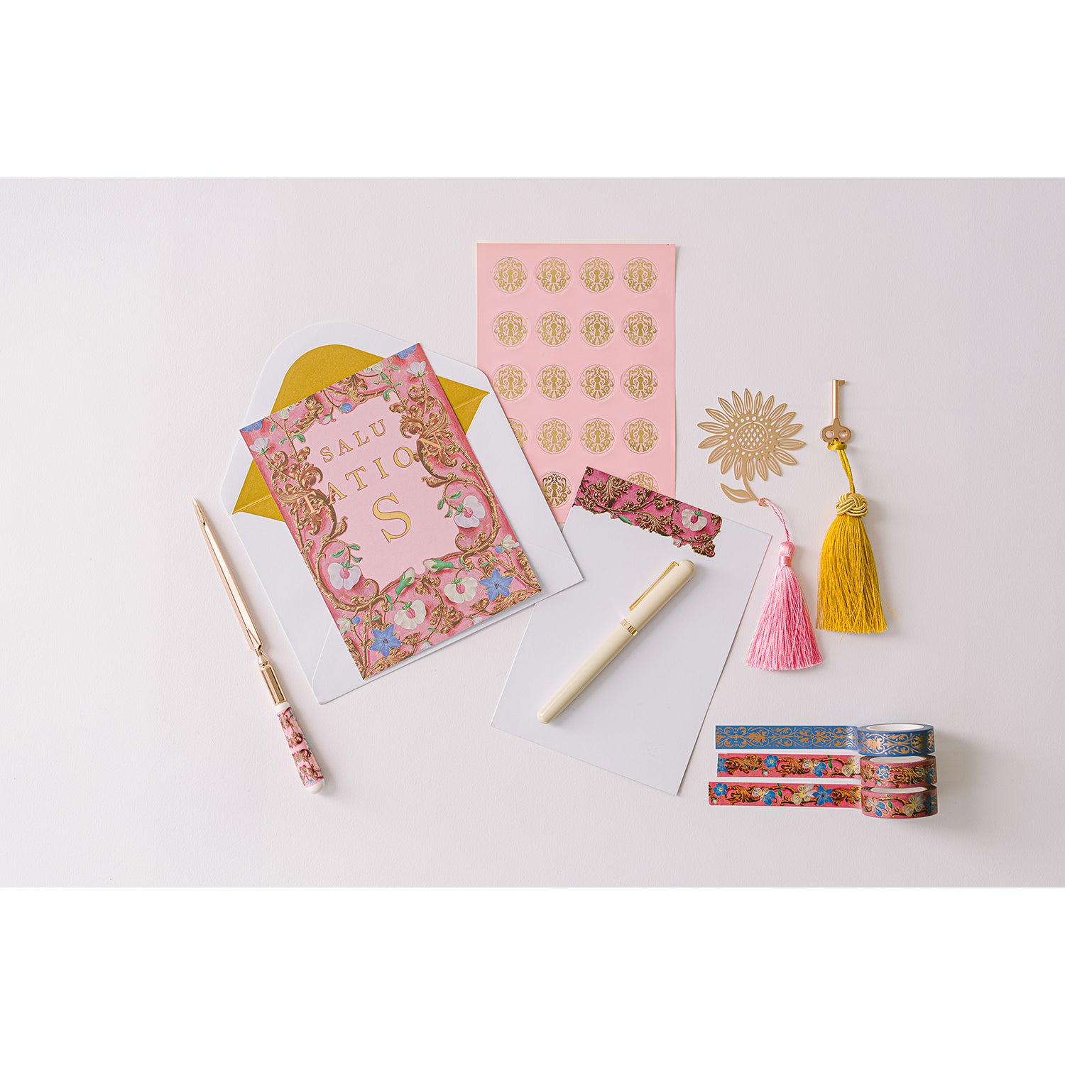 A pink lacquer keepsake box with a velvet interior, containing stationery items including writing sheets, metallic lined envelopes, stickers, washi tape, a metal bookmark with a tassel, and a ceramic letter opener. The box is open and displays the items arranged neatly.