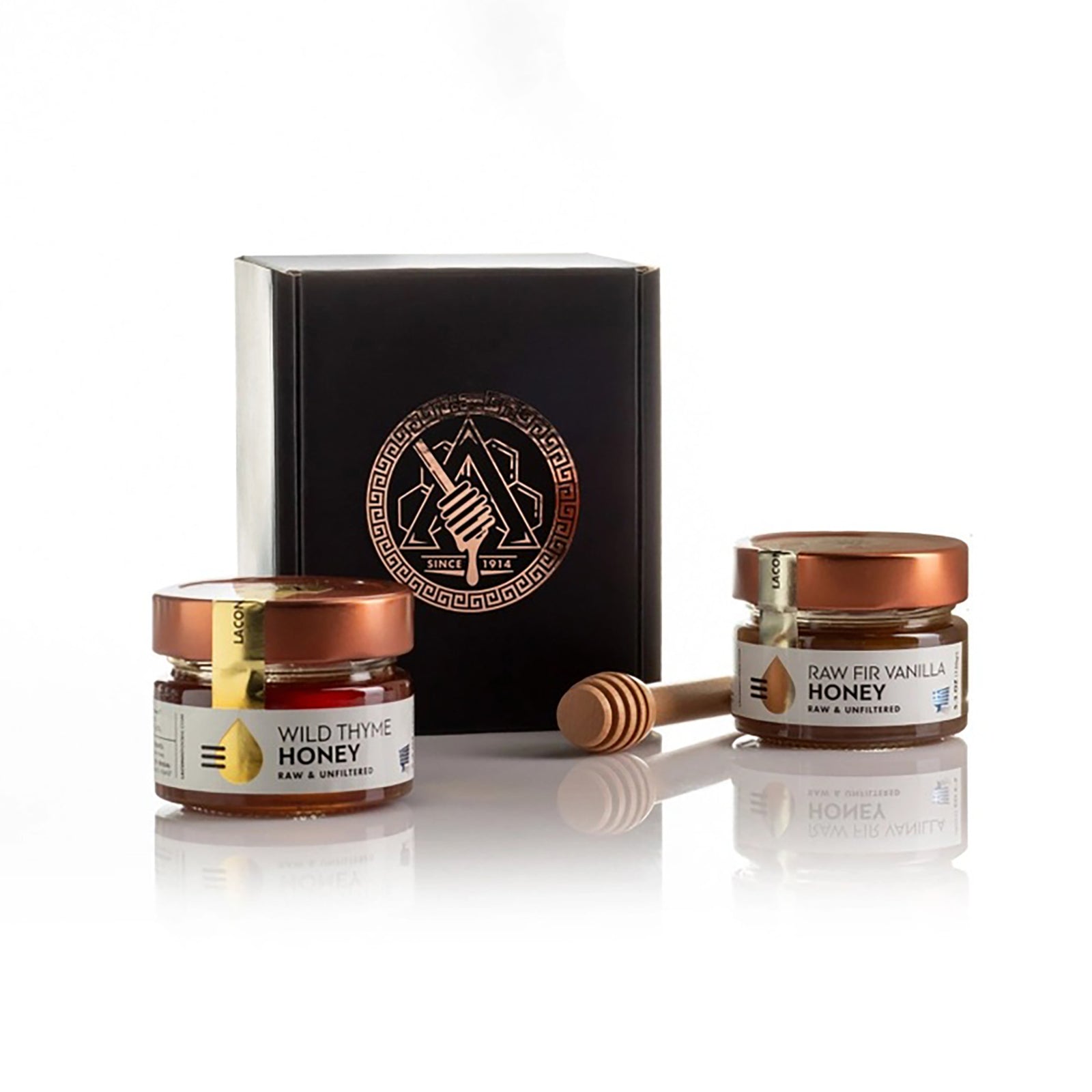 A honey gift set featuring two glass jars of honey with copper lids, a wooden honey dipper, and a black presentation box with a metallic circular logo, all on a reflective white surface.