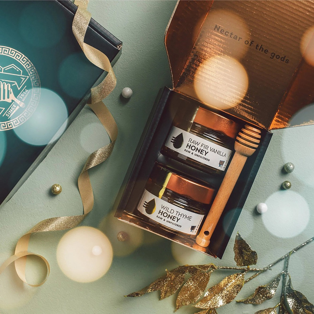 An open gift box containing two jars of honey and a wooden honey dipper, styled on a sage green surface with gold ribbon, decorative leaves, and soft glowing orbs. The jars are labeled Raw Fir Vanilla Honey and Wild Thyme Honey.