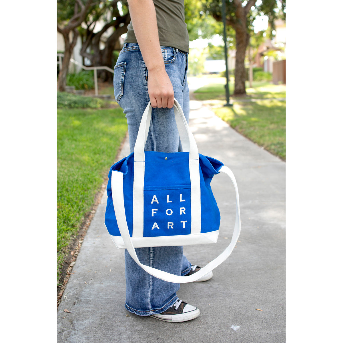 A person outdoors on a sidewalk holding a blue canvas tote bag with white handles and white side panels. The bag has the words 'ALL FOR ART' printed vertically in white on the front pocket.
