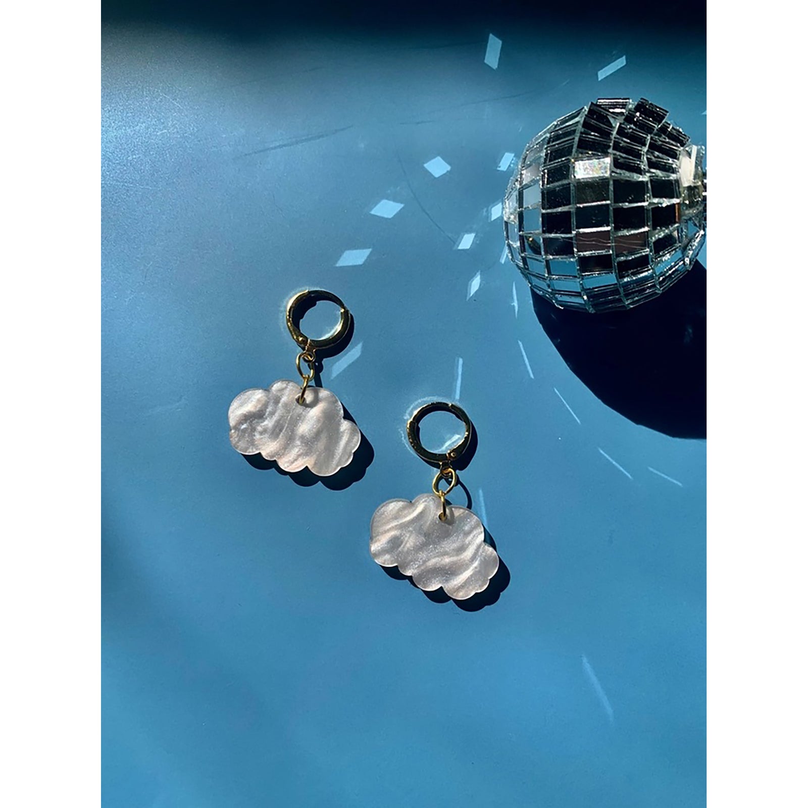 Pair of cloud-shaped earrings with gold hooks on a reflective blue surface with a disco ball.