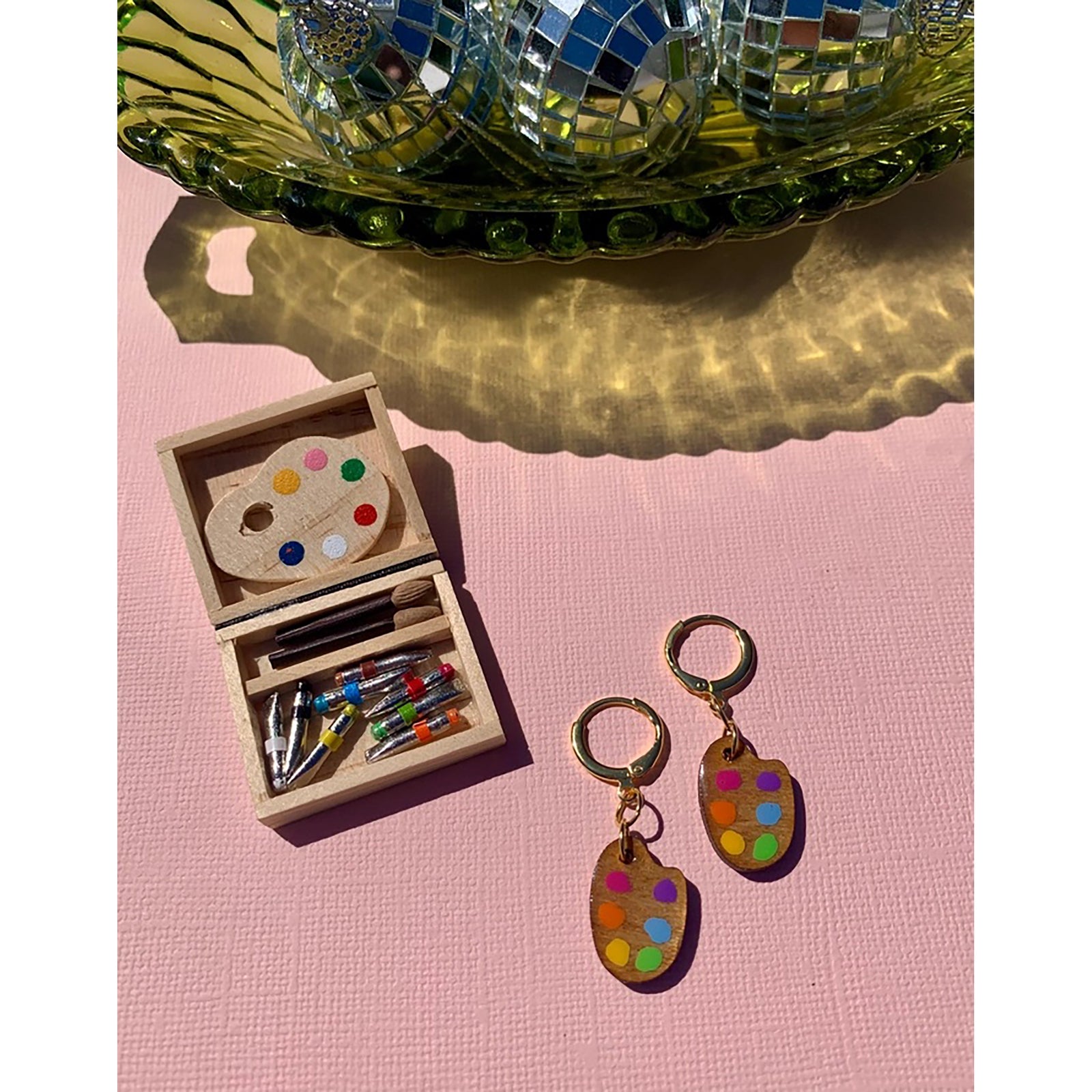 Colorful paint palette earrings on a pink surface with a small wooden box and disco balls in the background.