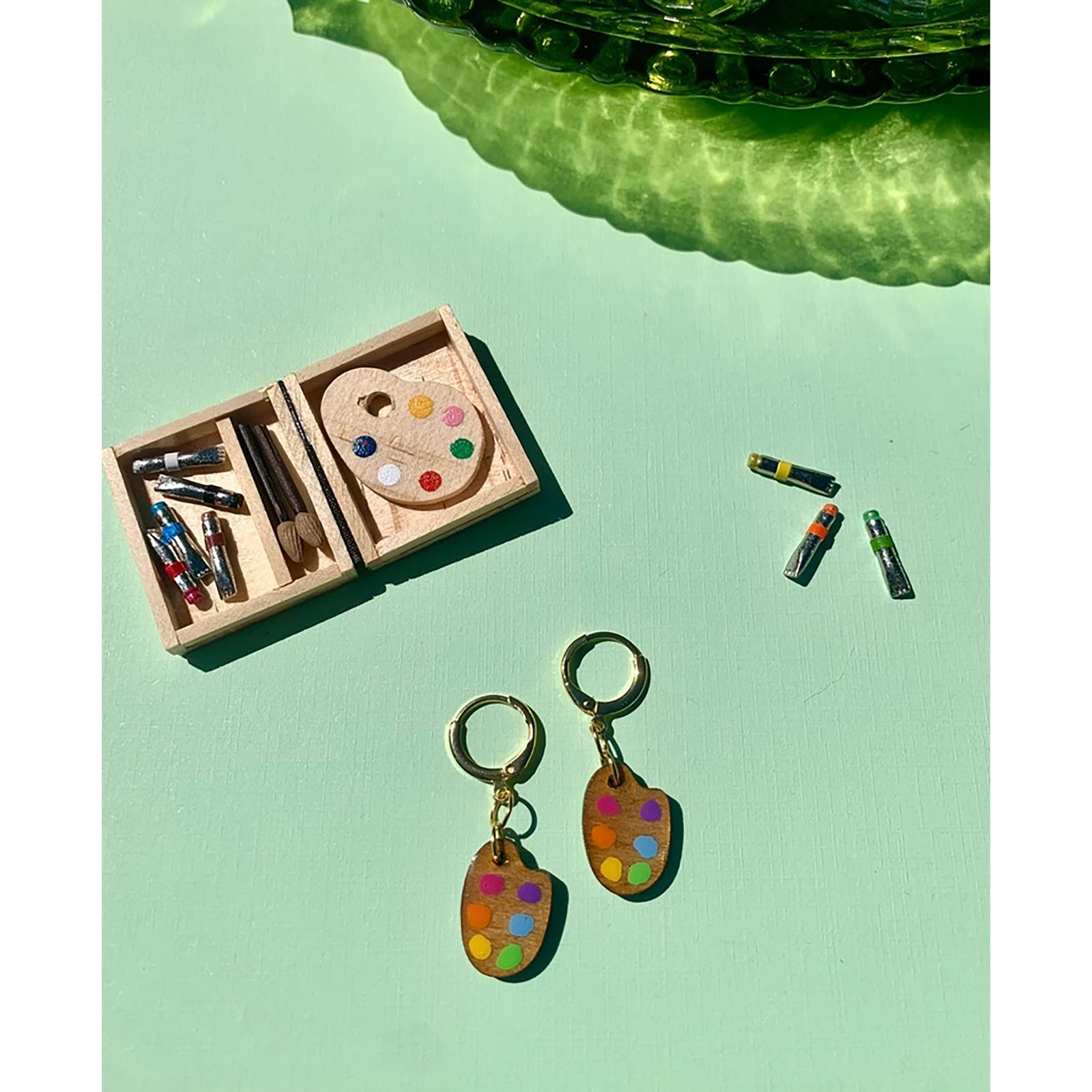 Colorful paint palette earrings on a pink surface with a small wooden box and disco balls in the background.