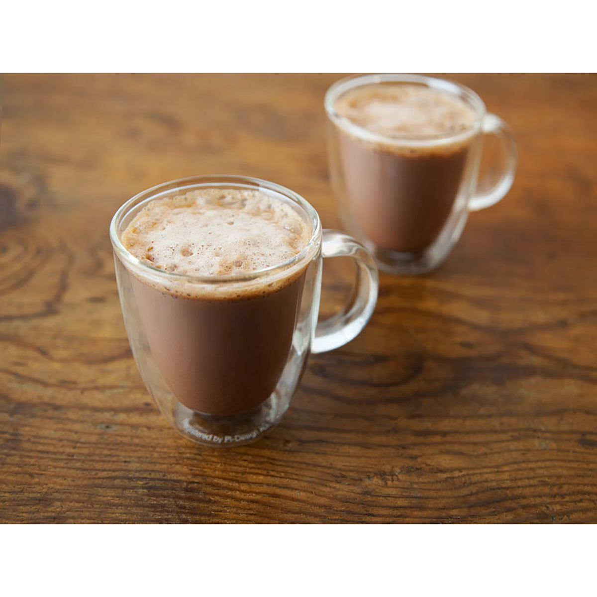 Two clear double-walled glass mugs filled with a frothy brown beverage, placed on a wooden tabletop.