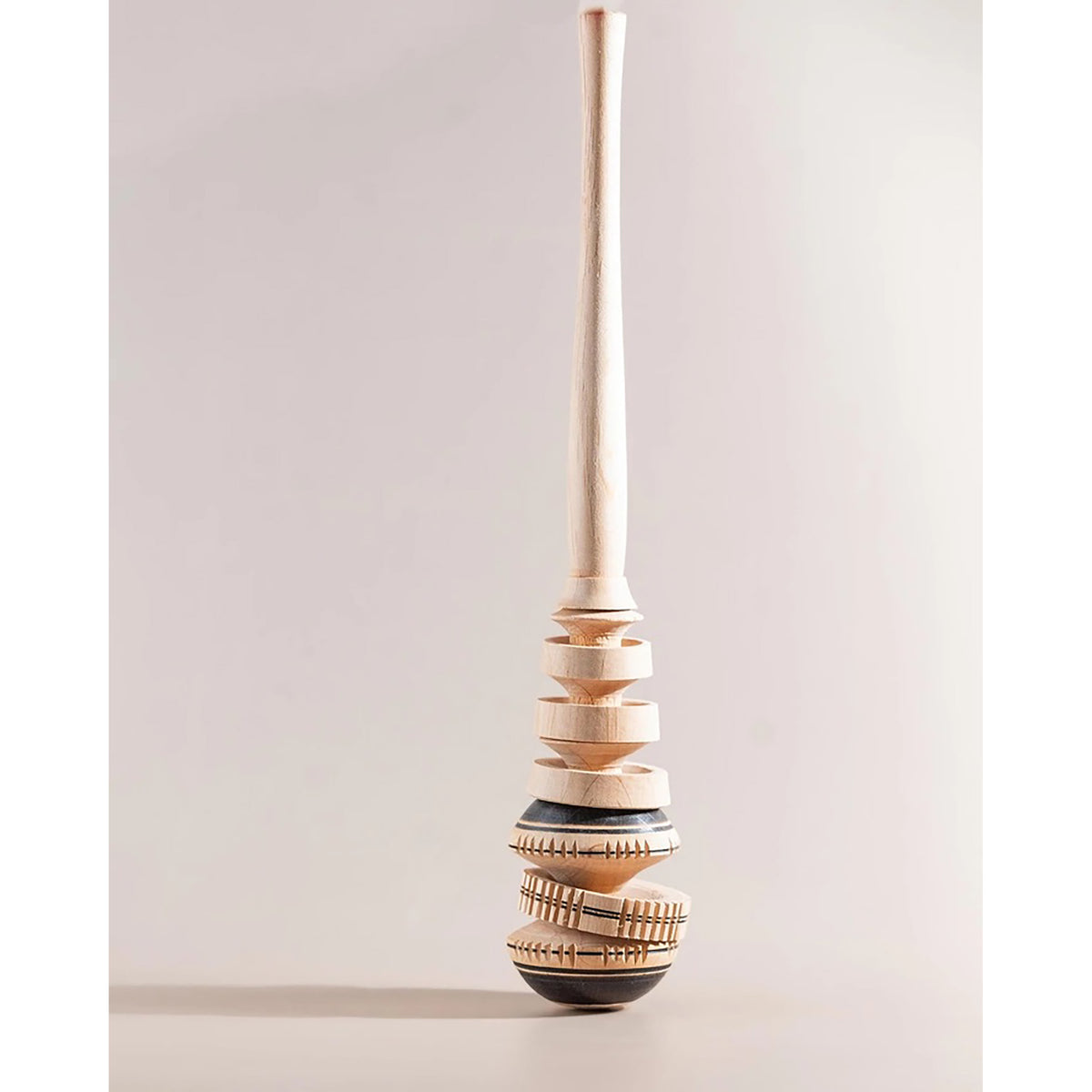 A tall, sculptural wooden whisk like item with a light natural wood finish and dark accents, photographed against a neutral light gray background.