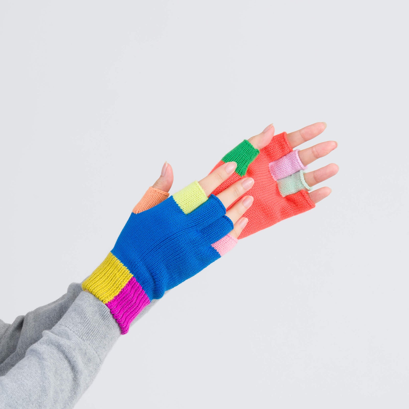 A pair of colorful fingerless knit gloves with a ribbed cuff. One glove is primarily blue with color blocks in pink and green, while the other is primarily red with color blocks in blue and yellow.