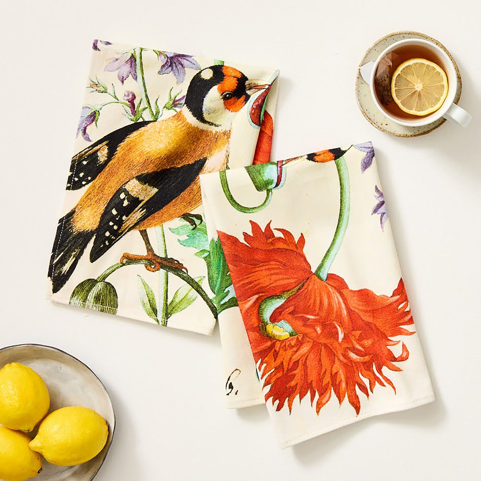 A tea towel with an illustration of a bird and a red flower, with green leaves on a white background.